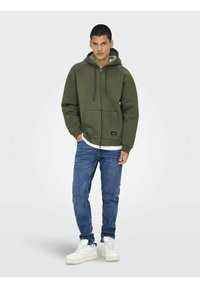 Grön zip-up hoodie med fodrad huva, framfickor och justerbara snoddar, som är ihopkopplad med blå jeans och vita sneakers.