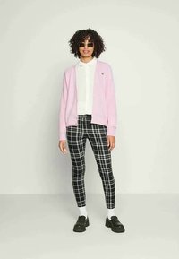 Cardigã rosa claro sobre uma camisa branca, combinado com leggings de xadrez preto e branco. Sapatos pretos e óculos de sol completam o look.