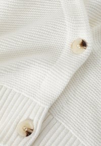 Cardigan bianco lavorato a maglia con texture a coste, due grandi bottoni rotondi in crema con accenti scuri e un aspetto morbido e accogliente.