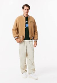 Pruun fliisist bomber-jakett ribadega mansettides, roheline kudumiga sviiter sinise mustriga, beežid cargo-püksid ja valged sneakerid.