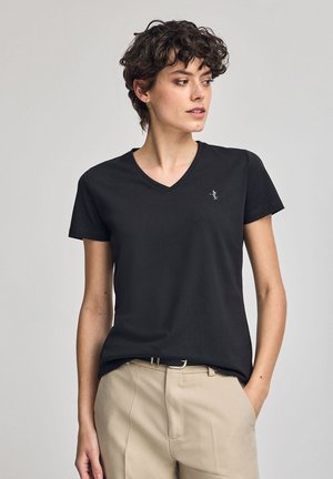 T-shirt nera a scollatura a V realizzata in tessuto morbido, con maniche corte e un logo discreto sul petto sinistro, abbinata a pantaloni beige.
