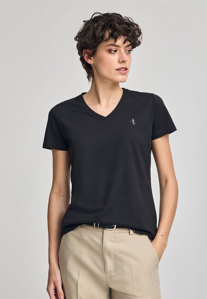 Czarna koszulka polo z dekoltem w kształcie litery V, wykonana z miękkiego materiału, z krótkimi rękawami i subtelnym logo po lewej stronie klatki piersiowej, w połączeniu z beżowymi spodniami.