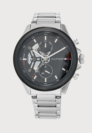Montre-bracelet Tommy Hilfiger argentée avec un bracelet à maillons en métal, un cadran foncé, des engrenages apparents et trois sous-cadrans.