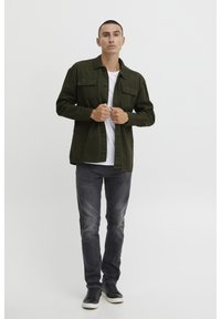 Giacca di cotone verde oliva con due tasche frontali, chiusura a bottoni, indossata sopra una t-shirt bianca, abbinata a jeans grigi e sneaker nere.