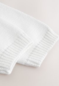 Manches d'un pull blanc en tricot avec des poignets côtelés posées à plat sur une surface claire, montrant la texture et les détails des mailles.