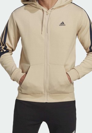Beżowa bluza Adidas na zamek z czarnymi paskami na rękawach, kieszeniami z przodu i logo Adidas na piersi, noszona z czarnymi spodniami.