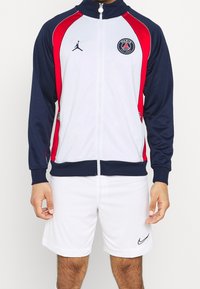 Chaqueta deportiva en blanco, azul marino y rojo. Presenta cierre de cremallera, puños acanalados y detalles con el logo. Combinada con pantalones cortos blancos y acentos de logo negros.