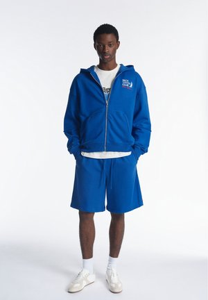 Personne debout portant un sweat à capuche bleu zippé avec le texte "Nice Yacht Club", un short bleu assorti, un t-shirt blanc, des chaussettes blanches et des baskets blanches.