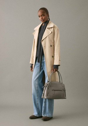 Donna che indossa un trench beige, un maglione scuro, jeans a gamba larga e scarpe marroni, che tiene una borsa grigia contro uno sfondo grigio semplice.