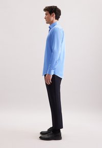 Camicetta azzurro chiaro a maniche lunghe con colletto a punta, texture liscia e dettagli con bottoni, abbinata a pantaloni scuri e scarpe nere.