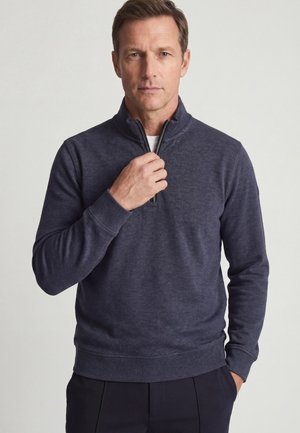 Man past de ritssluiting van een donkergrijze trui met kwartritssluiting aan, staande tegen een effen lichte achtergrond.