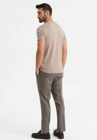 Beige bavlněné tričko s krátkými rukávy, spárované se světle šedými kalhotami a hnědými mokasínami. Model je vyfocen zezadu, mírně otočený doleva.