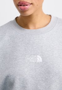 Světle šedá mikina vyrobená z měkké látky, s bílým vyšitým logem "The North Face" na hrudi. Jednoduchý design s klasickým kulatým výstřihem.