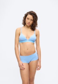 Νεαρή γυναίκα με σγουρά μαλλιά που φοράει ανοιχτό γαλάζιο μπραλετί και ταιριαστό σορτς, στέκεται μπροστά σε ένα απλό λευκό φόντο.