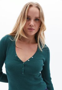 Jersey de manga larga en tono verde azulado con escote en V, que presenta tres botones en la parte delantera y un diseño ajustado. Textura suave, corte estilizado.