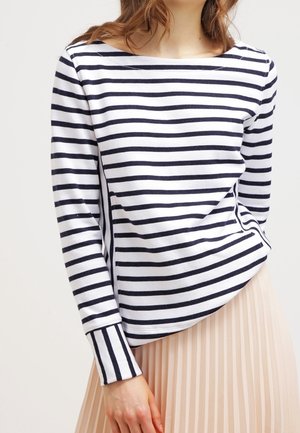 Kobieta w białym długim rękawie z czarnymi poziomymi paskami i jasnobeżową plisowaną spódnicą, stojąca na jednolitym tle.