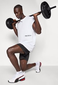 Barre noire portée sur les épaules par une personne portant un débardeur blanc avec le texte "PUMA CREW 48" et un short noir, avec des baskets blanches et rouges.
