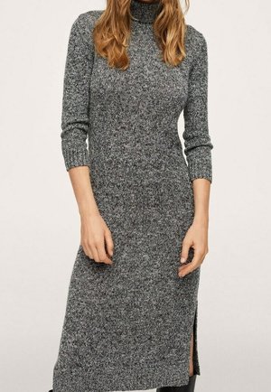 Kobieta ubrana w dopasowaną, szarą, dzianinową sukienkę midi z golfem, długimi rękawami i rozcięciem z boku oraz czarne botki.