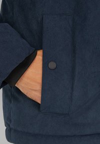 Dunkelblaues Cord-Jackenddetail mit einer strukturierten Oberfläche, ausgestattet mit einer Seitenasche mit einem runden schwarzen Knopf und einem gerippten Bündchen.