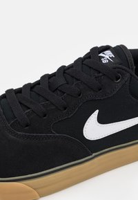 Zapatilla negra con parte superior de ante y tela, con un swoosh blanco de Nike, cordones negros y suela de goma color miel.