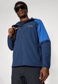 Veste coupe-vent bleu marine avec une touche de bleu sur les épaules, fermeture éclair et capuche avec cordon de serrage. Légère, avec des poches latérales et des poignets élastiques.