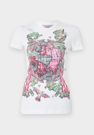 Fiatal nő fehér, grafikával díszített pólót visel piros és zöld mintával, fekete zsebes nadrágot cipzárokkal és csatokkal, fekete cipőt és fekete sapkát.