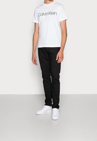 Man wearing white Calvin Klein T-shirt, black pants, and white sneakers standing against plain white background.