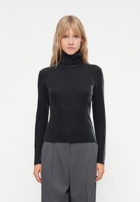 Μαύρο σουέτερ με ψηλό λαιμό σε κομψό, γυαλιστερό υλικό, με μακριά μανίκια και εφαρμοστό σχέδιο, συνδυασμένο με γκρι παντελόνι με φαρδιά ποδιά.