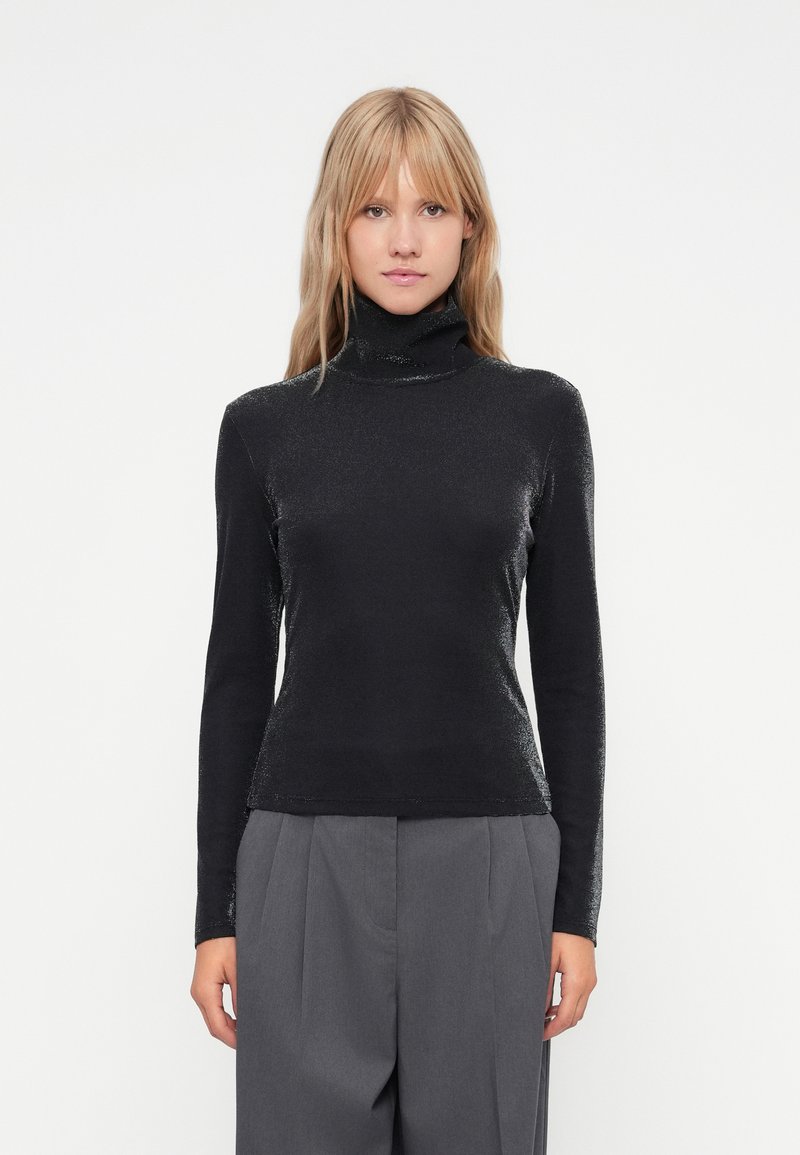 Μαύρο σουέτερ με ψηλό λαιμό σε κομψό, γυαλιστερό υλικό, με μακριά μανίκια και εφαρμοστό σχέδιο, συνδυασμένο με γκρι παντελόνι με φαρδιά ποδιά.