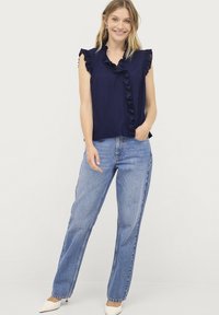 Marineblauw mouwloos topje met ruches en een voorpaneel detail, gecombineerd met lichtblauwe rechtlopende jeans en crèmekleurige puntige schoenen.
