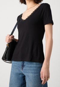 Maglietta nera a maniche corte con scollatura a V in pizzo ondulato, abbinata a jeans in denim blu e a una borsa nera con dettagli in hardware dorato.