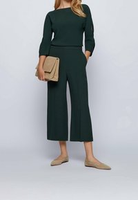 Mörkgrön kort jumpsuit med trekvartsärmar, slät textur och båtformad halsringning; kombinerad med en beige clutch och nude ballerinas.