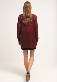 Lång burgundy kofta med strukturerad stickning, med diamantmönster, långa ärmar och sidslitsar, bärs över en svart klänning och klackskor.