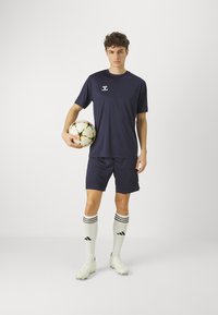 Tumesinine sportlik särk ja lühikesed püksid, koos valgete sokkidega, millel on mustad triibud. Modell hoiab mitmevärvilist jalgpalli.