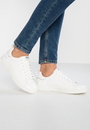 Person trägt weiße Schnürschuhe und blaue Jeans und steht mit einem leicht angehobenen Fuß auf einer hellgrauen Oberfläche.