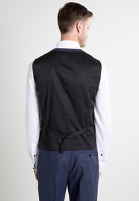 Schwarze Weste mit hohem Rücken, stoffgebundenem Verschluss, getragen über einem weißen Hemd. Die Hose ist dunkelblau mit einer strukturierten Oberfläche.