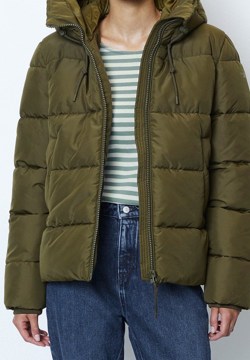 Doudoune verte olive avec capuche, comportant une fermeture éclair, des accents à cordon et une texture matelassée. Portée sur un haut rayé.