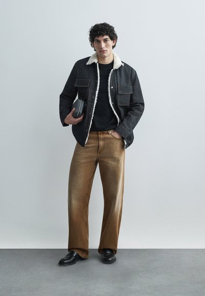 Džínová bunda s límcem z beránčí kožešiny, černé tričko a světle hnědé džíny s širokými nohavicemi. Drží malý černý doplněk, stojí na šedé podlaze.