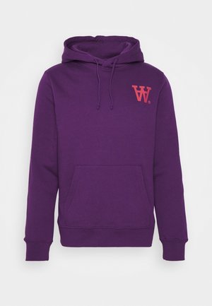 Henkilö, jolla on päällään violetti huppari, beiget housut, joissa on "Tommy"-logo, ja valkoiset lenkkarit, seisoo ulkona harmaalla päällystetyllä tiellä.