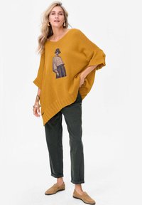 Musztardowe dziergane ponczo z asymetrycznym dołem i graficznym wzorem na przodzie, zestawione z ciemnozielonymi spodniami i beżowymi mokasynami.