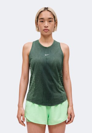 Femme aux cheveux blonds courts portant un haut sans manches à motifs vert foncé et un short de sport vert clair, debout devant un fond uni.