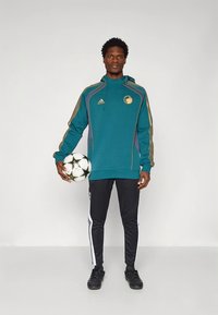 Tirkizna majica s kapuljačom zlatnih naglasaka i logotipom lava, uparena s crnim hlačama s bijelim bočnim prugama i crnim sportskim cipelama, drži nogometnu loptu.