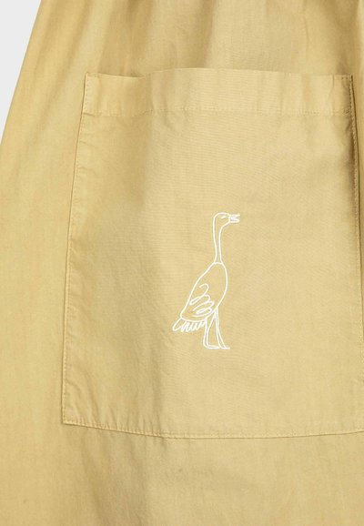 Poche en tissu beige avec un contour brodé blanc représentant une oie debout, cousue au centre de la poche.