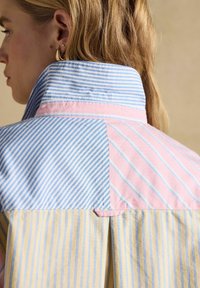 Svetlolaska ženska z visokim ovratnikom na srajci z mešanimi modrimi, roza in rumenimi navpičnimi in diagonalnimi črtami, vidnimi zlati obročki za ušesi.