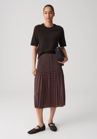 Pull marron à manches courtes, jupe maxi bordeaux à motifs avec des plis, et chaussures noires. Le mannequin tient un petit sac noir.