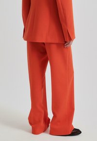 Oransje strukturert blazer og løstsittende bukser i et glatt stoff; buksene har brede ben og subtle plisser; kombinert med svart fottøy.