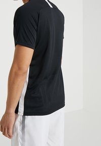 Persona que lleva una camiseta atlética negra de manga corta con paneles laterales blancos y pantalones cortos blancos con un detalle de rayas negras en el lateral.