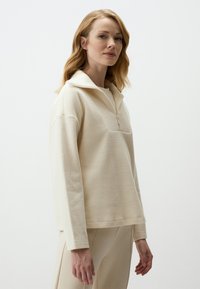 Cremfarbener Pullover aus strukturiertem Stoff, mit halbem Reißverschluss, großzügigem Schnitt und überschnittenen Schultern, kombiniert mit passenden Hosen.