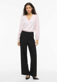 Blouse rose clair avec col en V et manches longues bouffantes, associée à un pantalon noir à jambes larges et des escarpins noirs à bouts pointus avec des accents dorés.