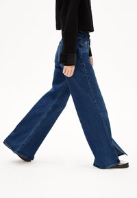 Wijde jeans in donkerblauwe denim met een onafgewerkt zoom en zijsplitten. Heeft een klassiek vijf-pocket ontwerp en riemlussen.